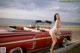 A woman leaning on the hood of a red convertible car.